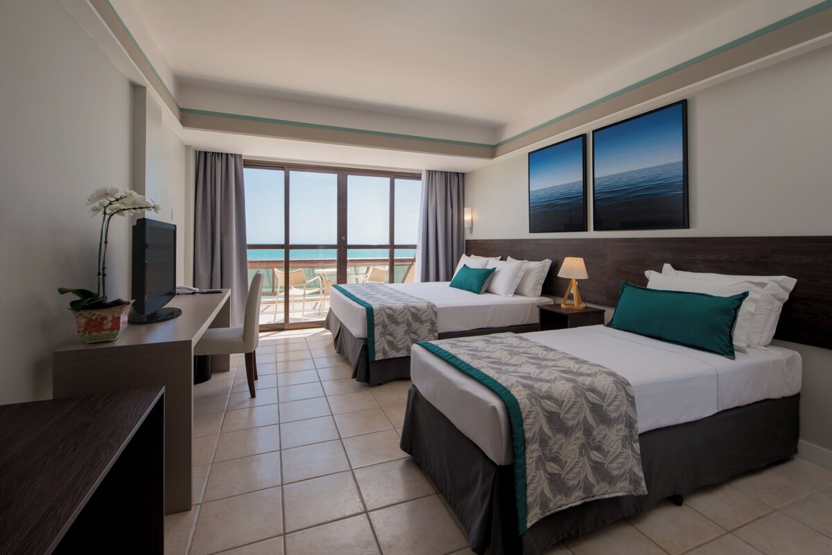 A hotel room with two beds, a desk, a TV, and a balcony with a view of the sea.