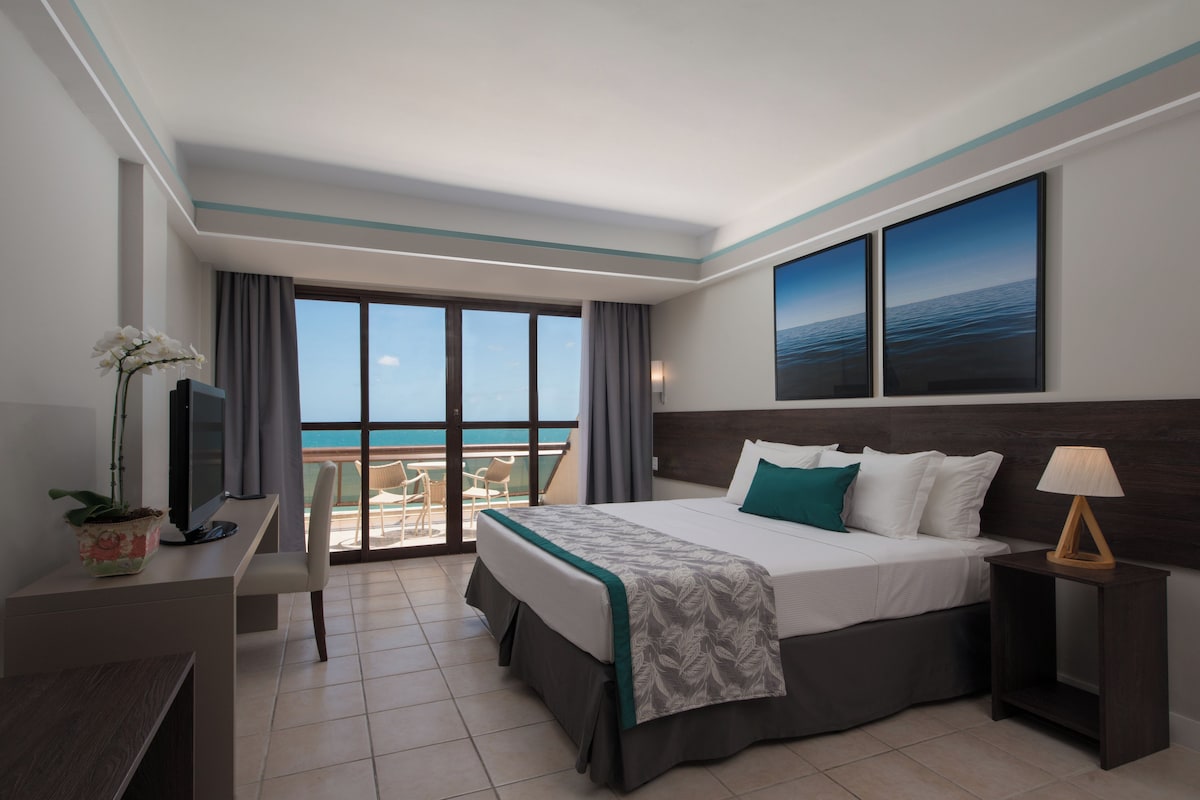Een hotelkamer met een bed, een bureau, een televisie, een raam met uitzicht op zee en een balkon.