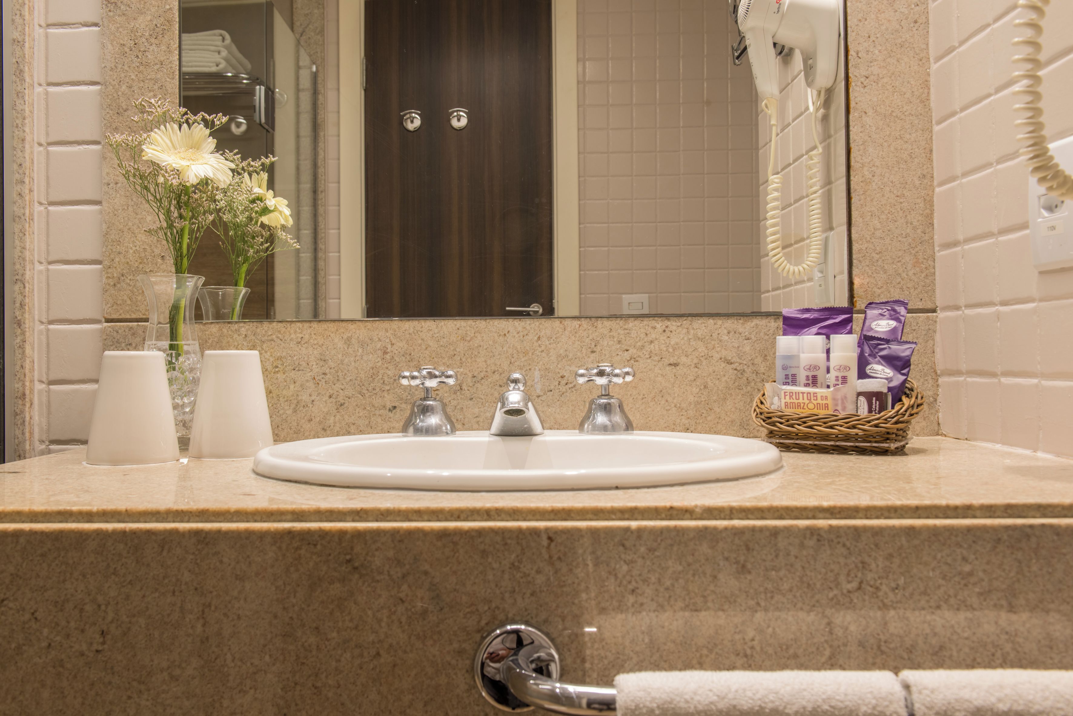 A bathroom with a sink, two faucets, a mirror, a hairdryer, a vase with flowers, and a basket with toiletries.