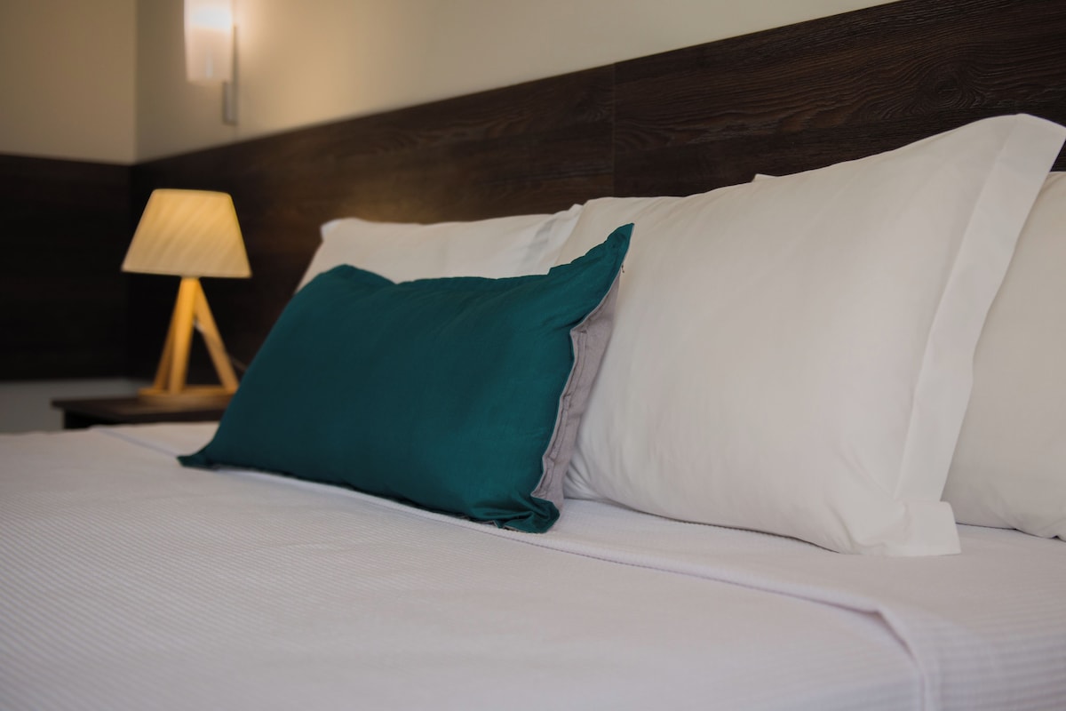 Een keurig opgemaakt bed met witte lakens en een turquoise kussen, een houten hoofdbord en een bedlampje.