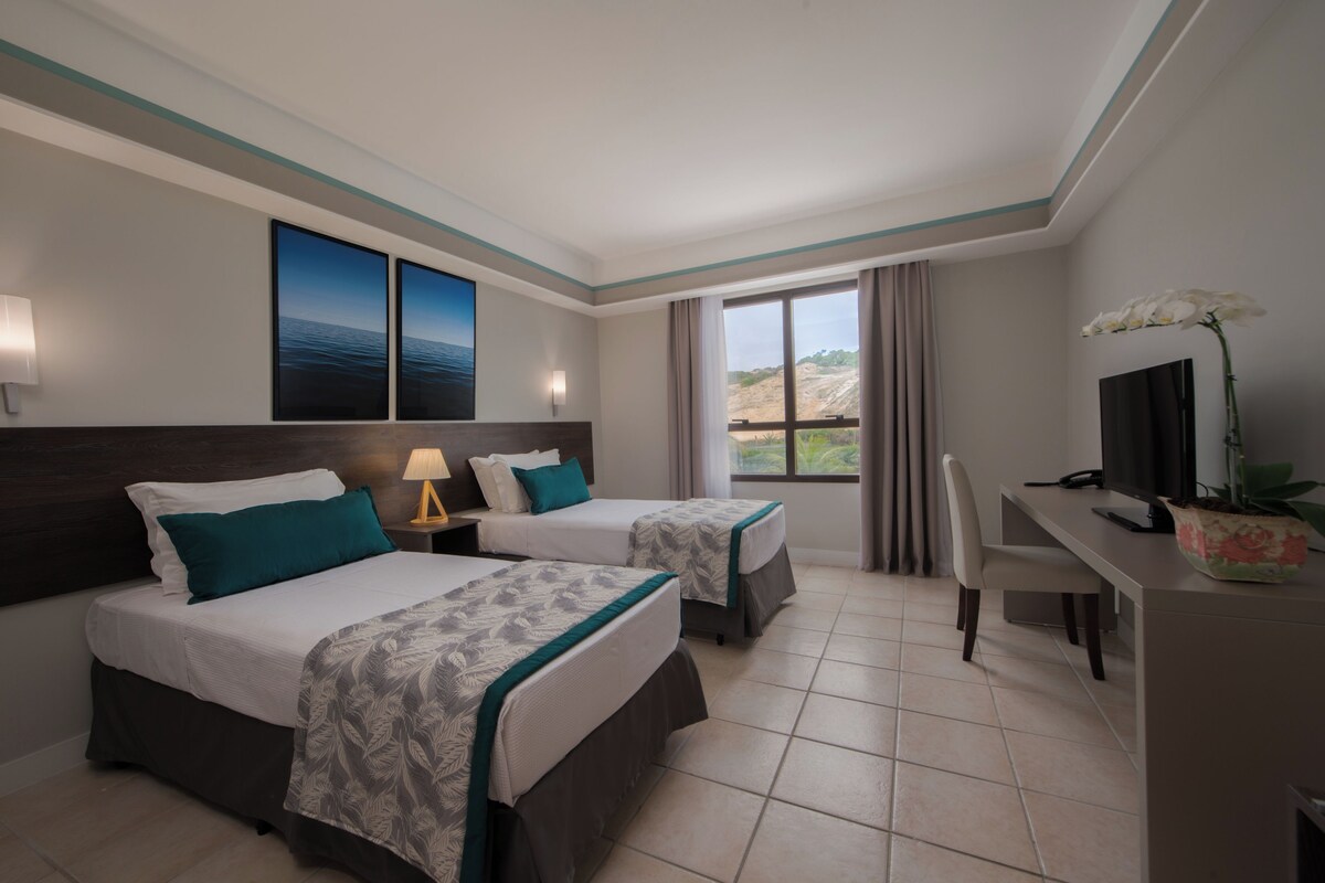 A hotel room with two beds, a desk, a television, and a view of the outdoors.