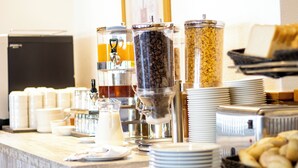 Disposição de buffet com diversos dispensadores de comida, incluindo café, cereais e outros itens, junto com pilhas de pratos e tigelas.