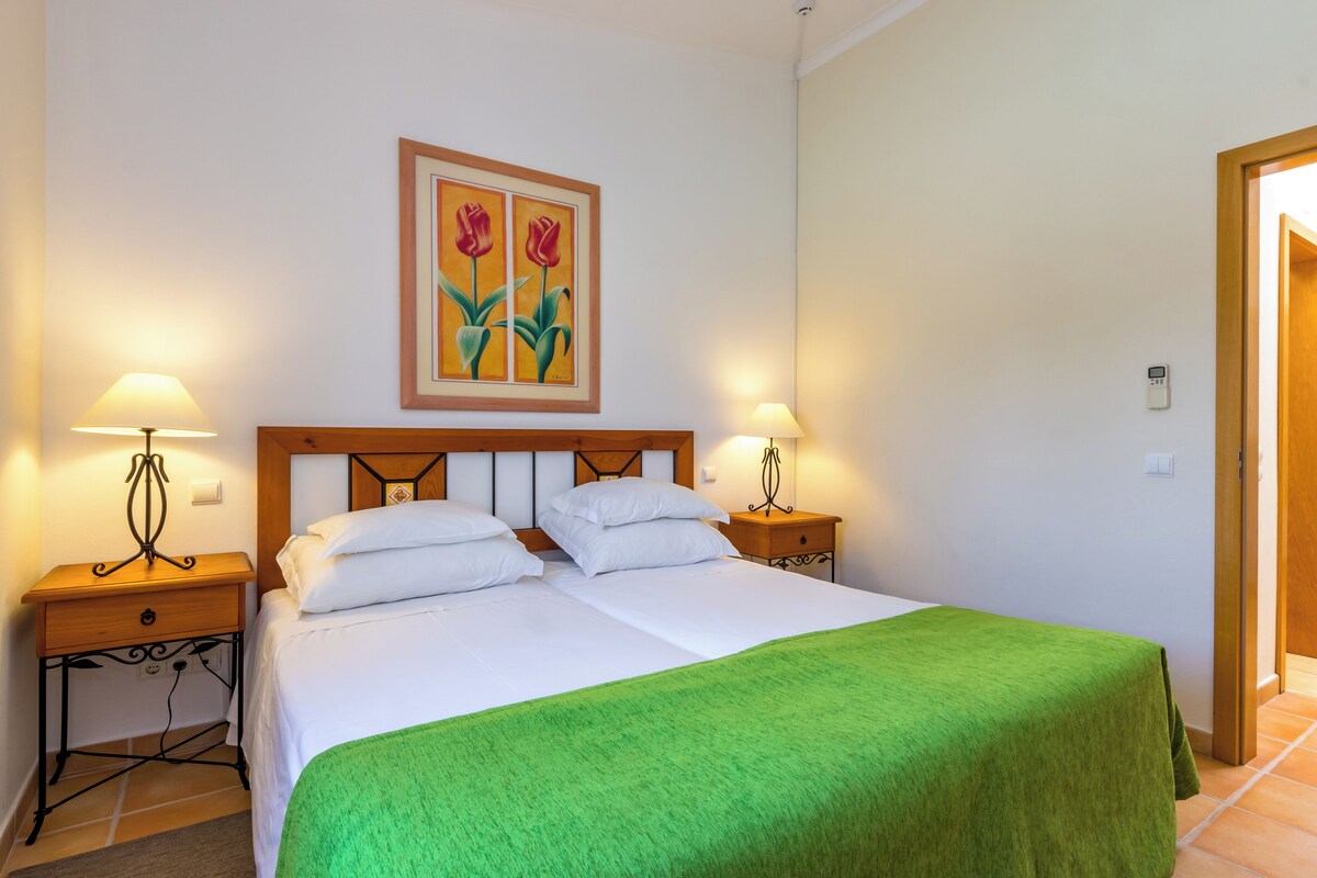 A hotel room with a double bed, two bedside tables with lamps, a green bedspread, and a painting of tulips on the wall.