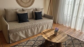 Uma sala de estar com um sofá bege, duas almofadas escuras, uma mesa de centro redonda com um vaso, um abajur e um painel de parede trançado.