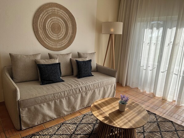 Un salón con sofá color beige, dos almohadas oscuras, una mesa de centro redonda con un florero, una lámpara de pie y un tapiz de pared tejido.