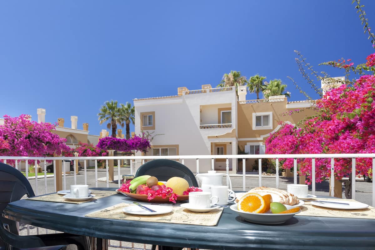 A table set with breakfast items, a white railing, and a building with balconies and pink flowers in the background.