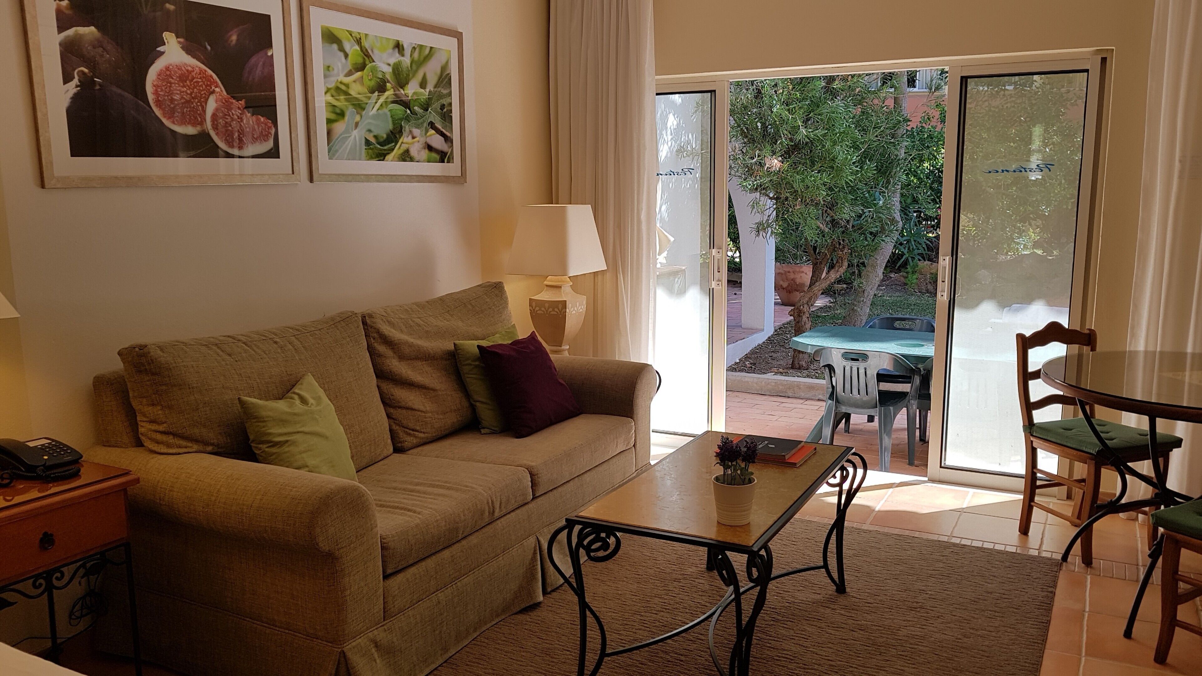 A living room with a sofa, a coffee table, and a dining area with chairs. There are two framed pictures on the wall.