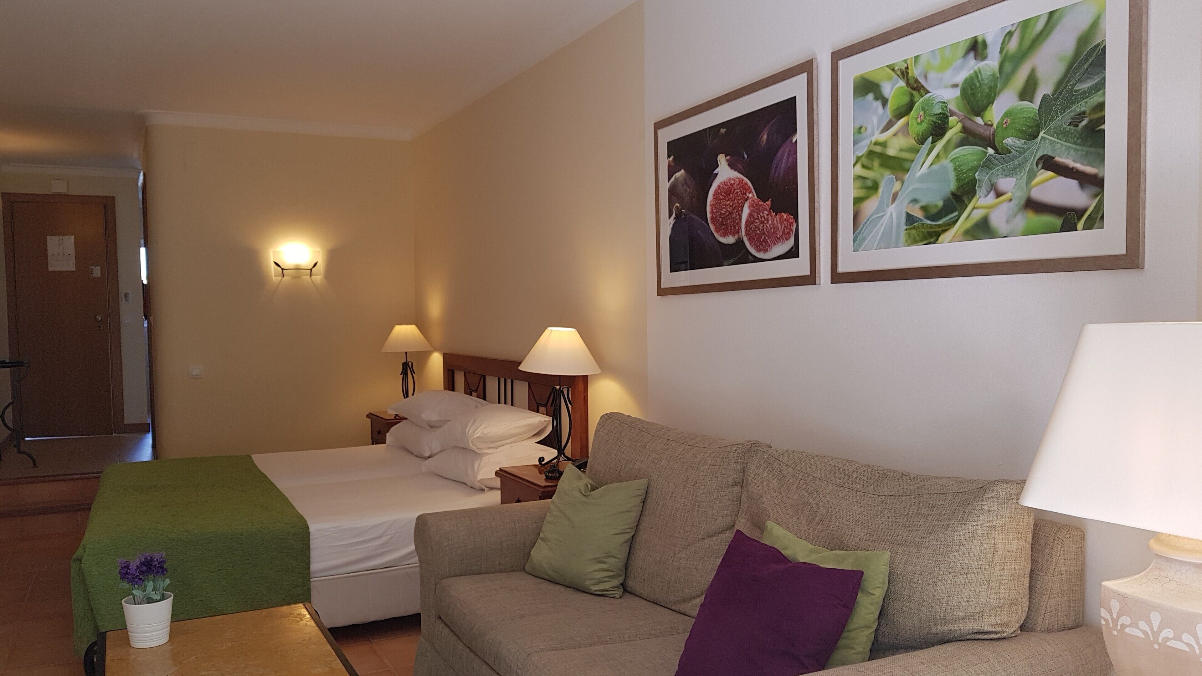 A hotel room with a bed, a sofa, two wall-mounted lamps, and two framed pictures of figs.