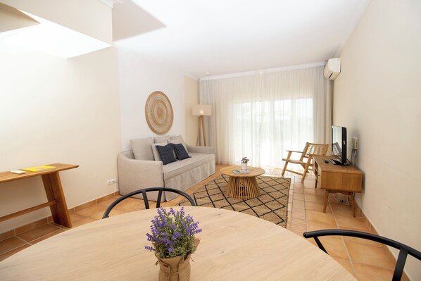 A modern living room with a sofa, a dining table with a vase of flowers, a coffee table, and a TV on a wooden stand. - Pestana Palm Gardens (Lagoa)