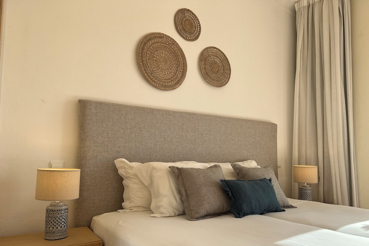 A neatly made bed with white and gray bedding, a textured headboard, and a bedside lamp on a wooden nightstand.