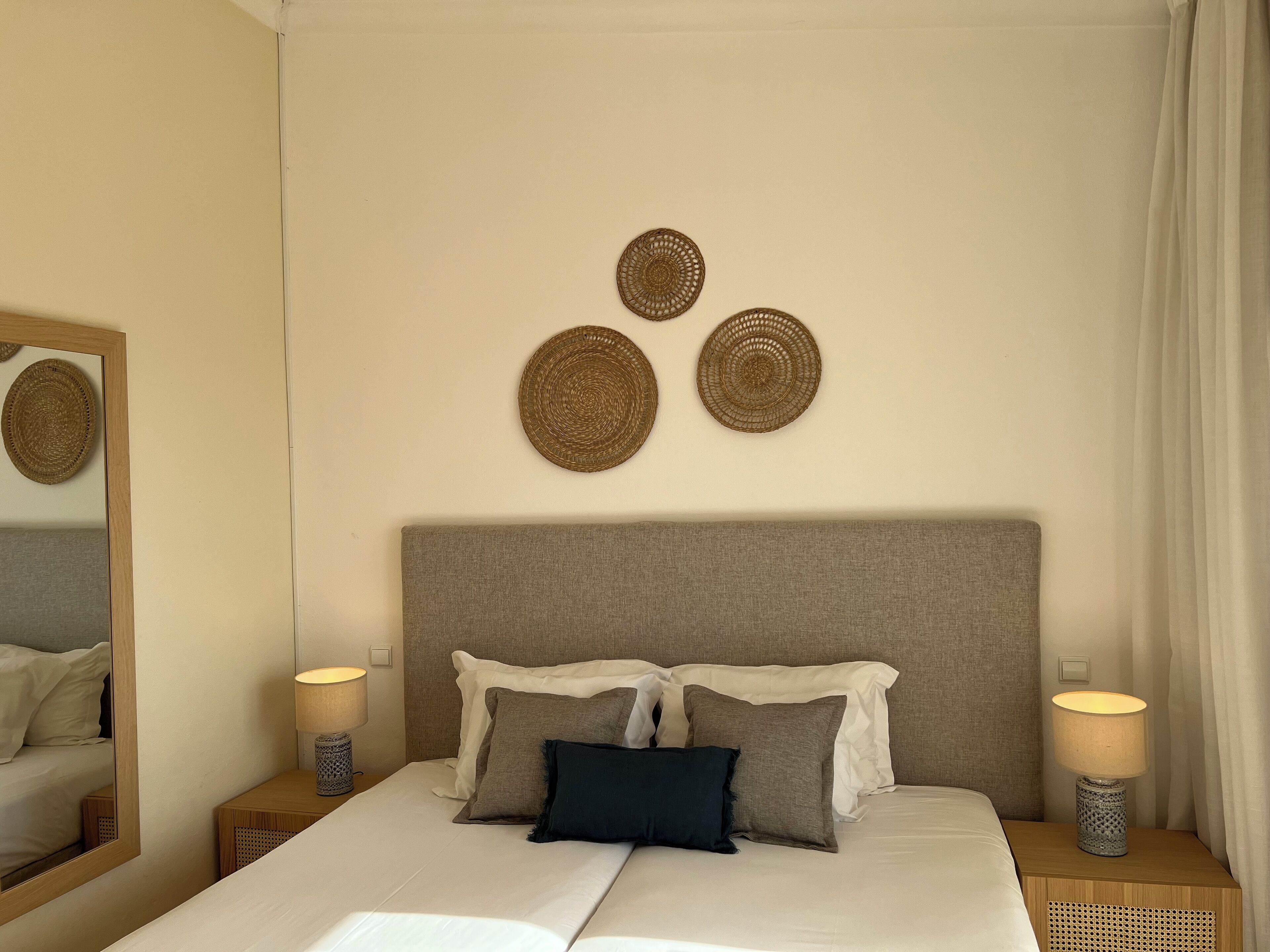 Una cama bien hecha con cabecera gris, dos lámparas de noche y tres piezas de arte mural tejidas.