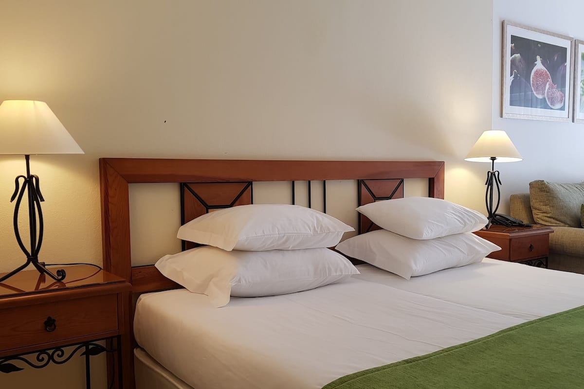 A hotel room with a wooden headboard, a bed with white linens, two bedside tables with lamps, a green throw blanket, and two framed pictures on the wall.