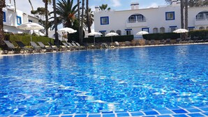 Uma piscina com água azul cristalina, cercada por espreguiçadeiras e guarda-sóis, e um edifício com paredes brancas e detalhes em azul ao fundo.