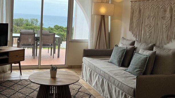 Sala de estar com sofá, mesa de centro, televisão e vista para o oceano através de grandes janelas.