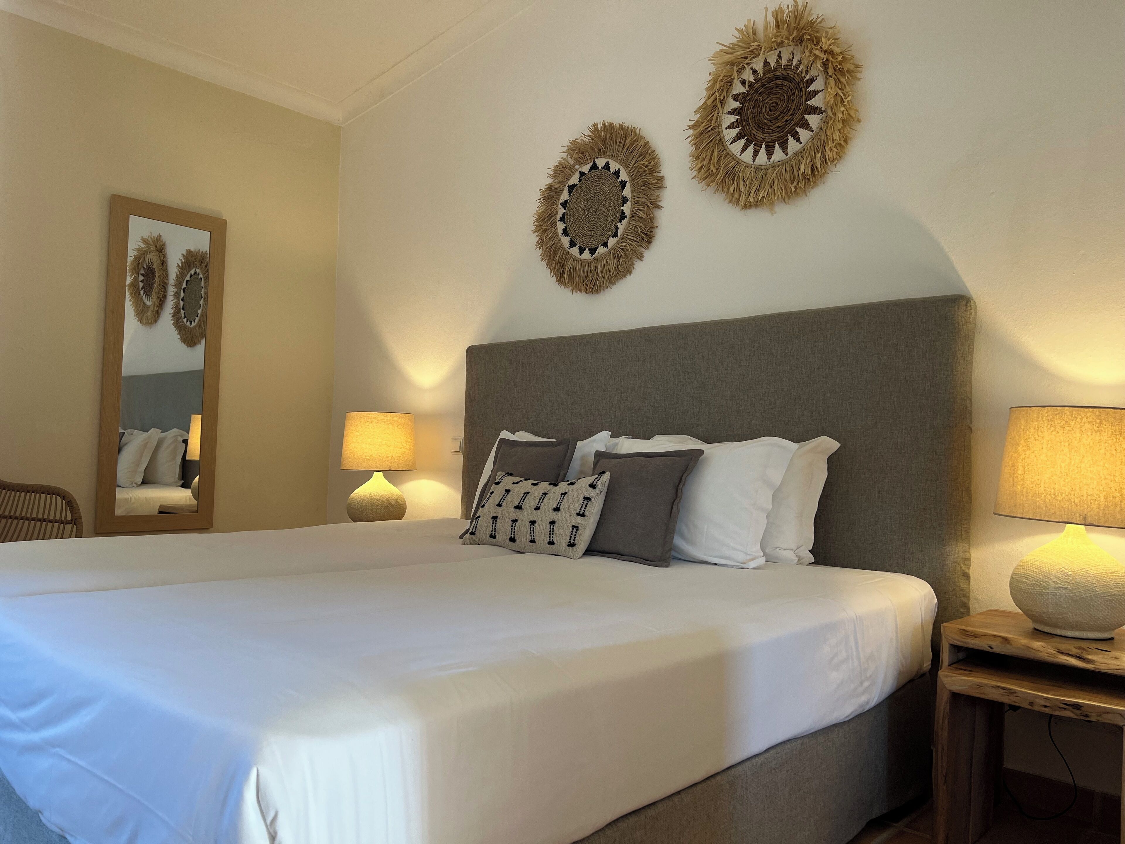 A large bed with white linens, a grey headboard, and multiple pillows. There are two wall-mounted lamps on either side of the bed.