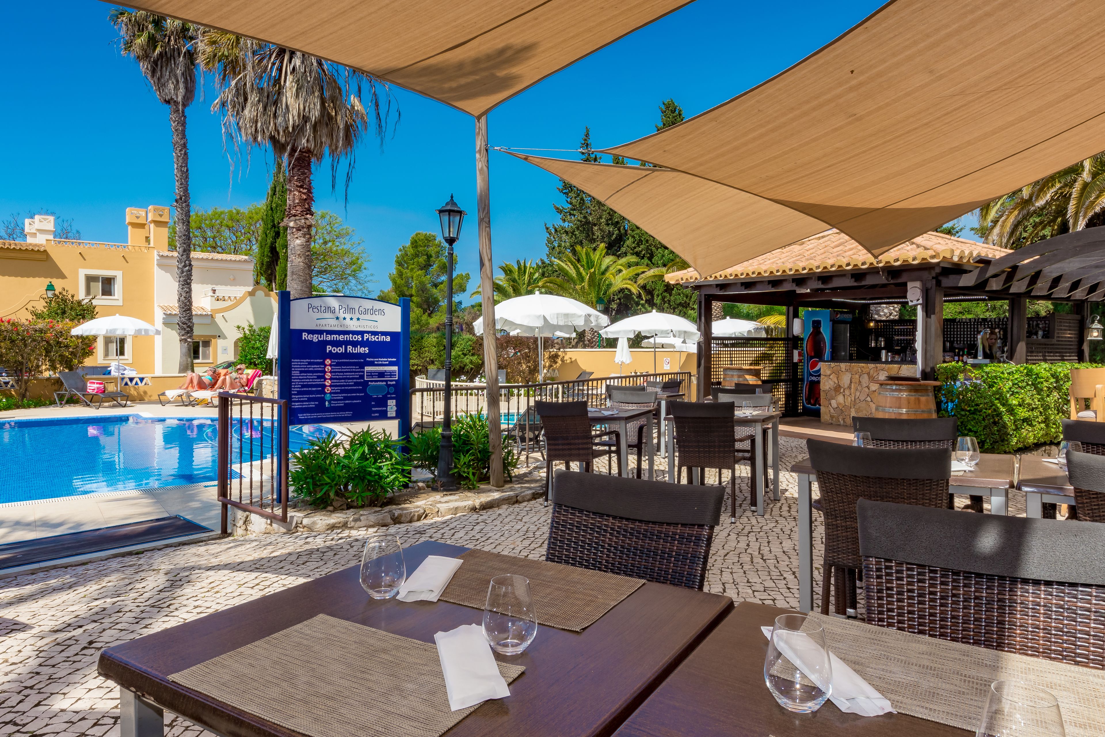 An outdoor pool area with lounge chairs and umbrellas, a covered seating area with tables and chairs, and a sign indicating pool rules.