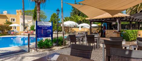 An outdoor pool area with lounge chairs and umbrellas, a covered seating area with tables and chairs, and a sign indicating pool rules.