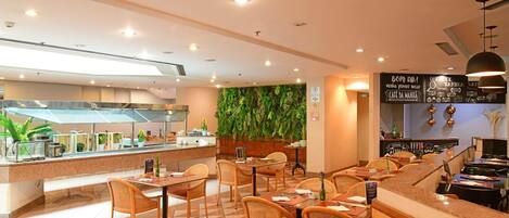 Un restaurante con zona de bufé, sillas de mimbre y una pared decorada con plantas.