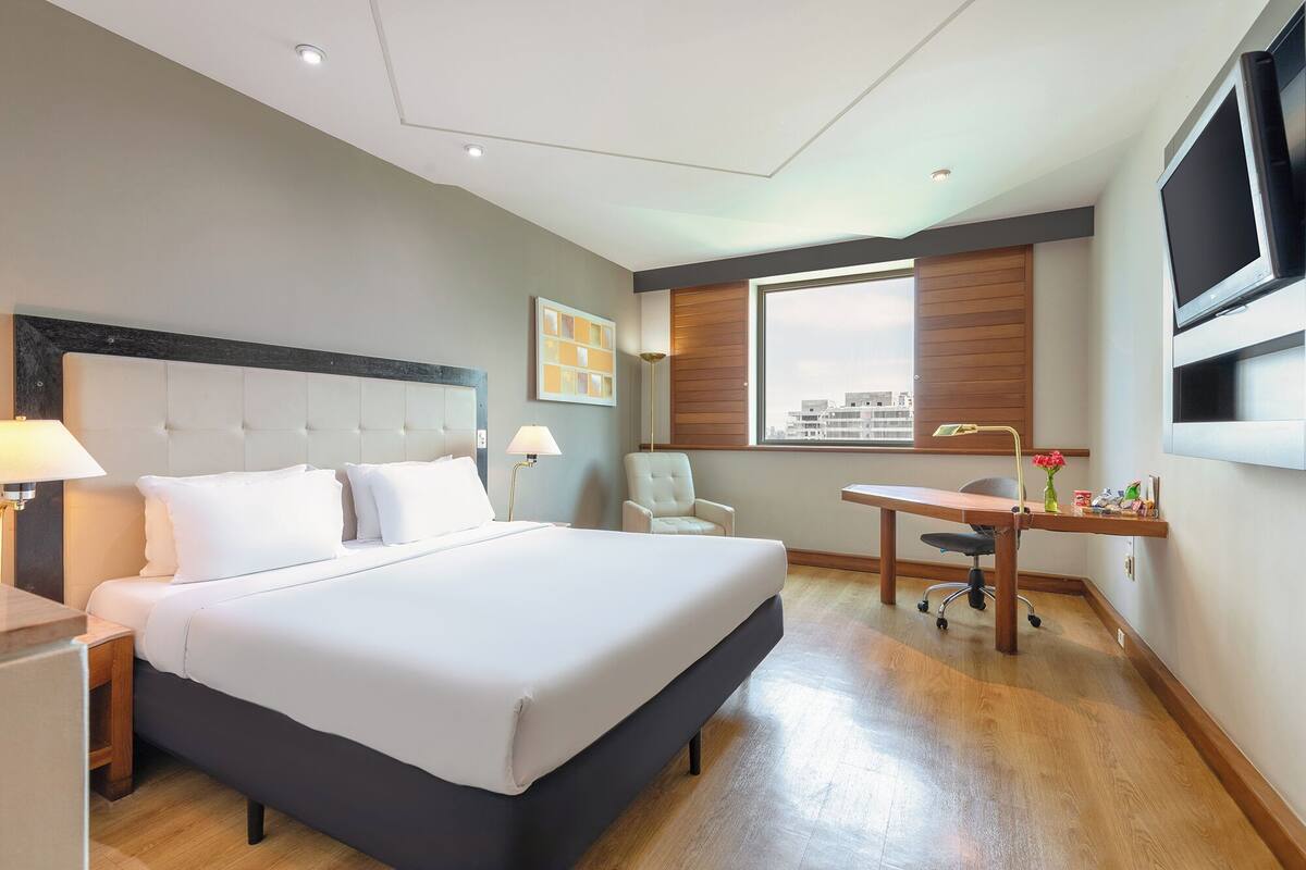Ein Hotelzimmer mit Bett, Schreibtisch, Stuhl, Lampe, Fernseher und einem Fenster mit Blick auf die Stadt.