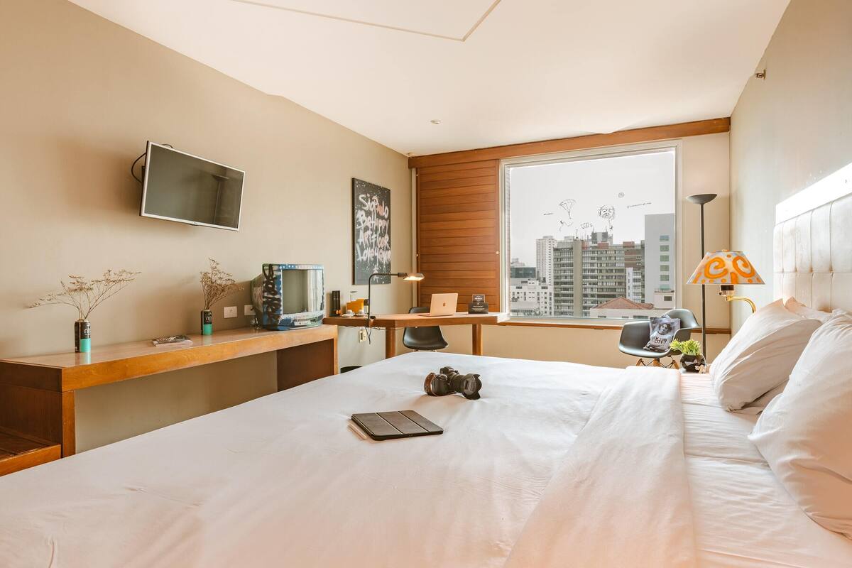 Ein Hotelzimmer mit Bett, Schreibtisch, Stuhl und einem großen Fenster mit Blick auf die Stadt.