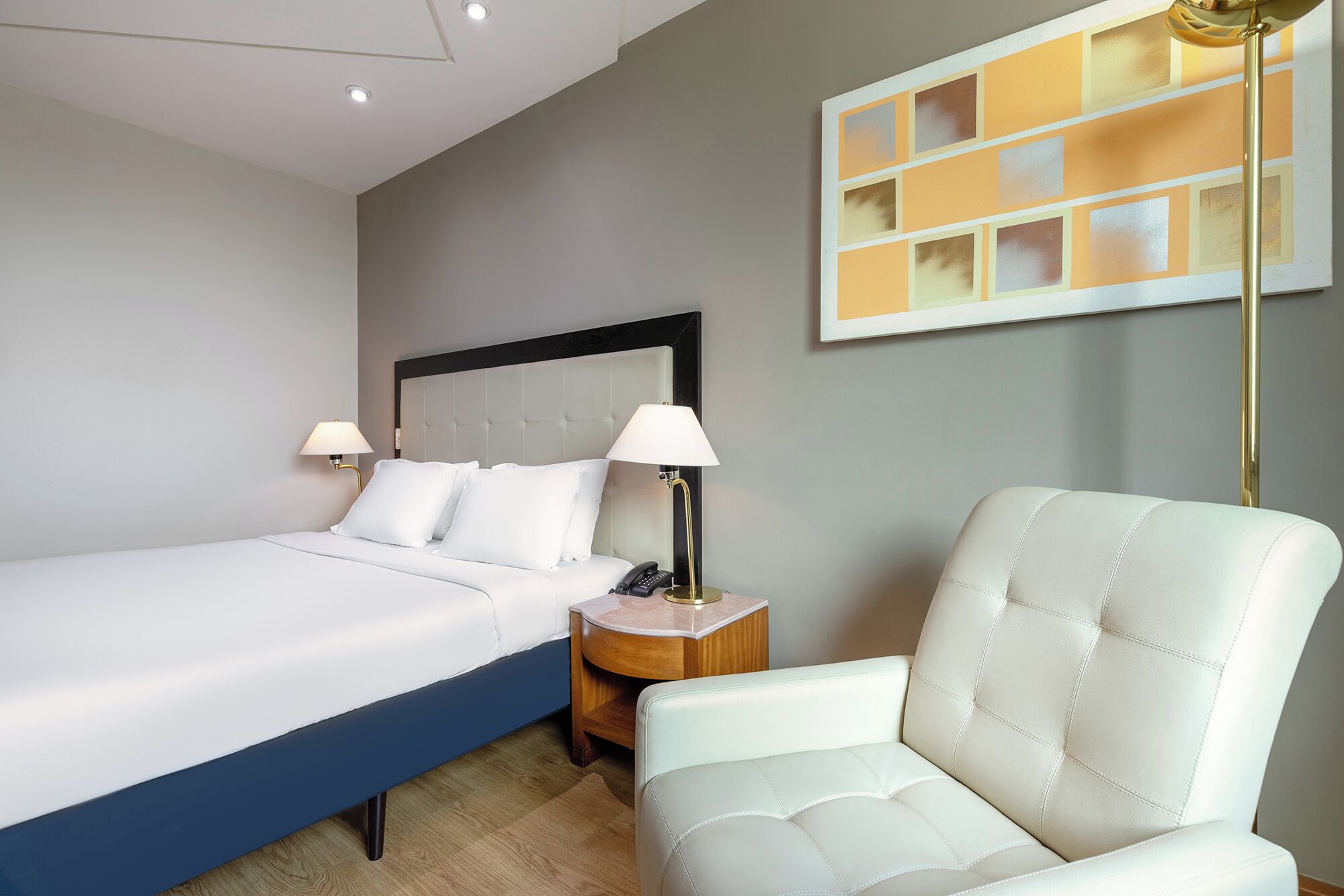 Camera d'albergo con un letto, abat-jour, comodino, una sedia e un quadro appeso al muro.