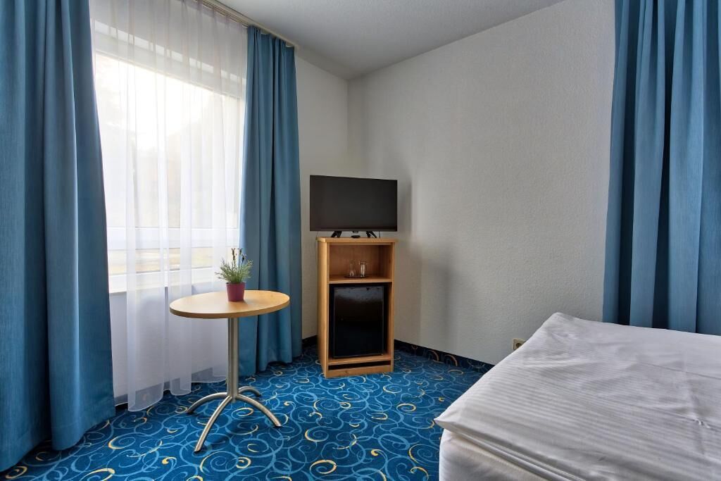 Et hotelværelse med en seng, et lille bord med en plante, et fjernsyn på et stativ og et vindue med blå gardiner.