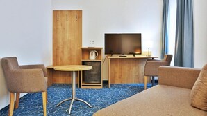 Ein Hotelzimmer mit einer Couch, zwei Sesseln, einem kleinen runden Tisch, einem Fernseher, einer Holzkommode und einer Kaffeemaschine.