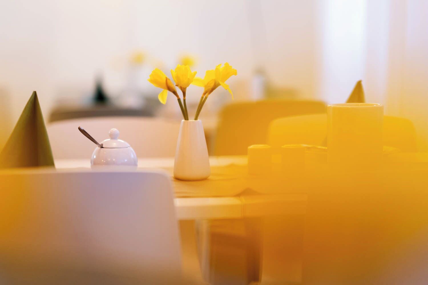 A table with a white vase containing yellow flowers, a small white container, and a yellow candle.