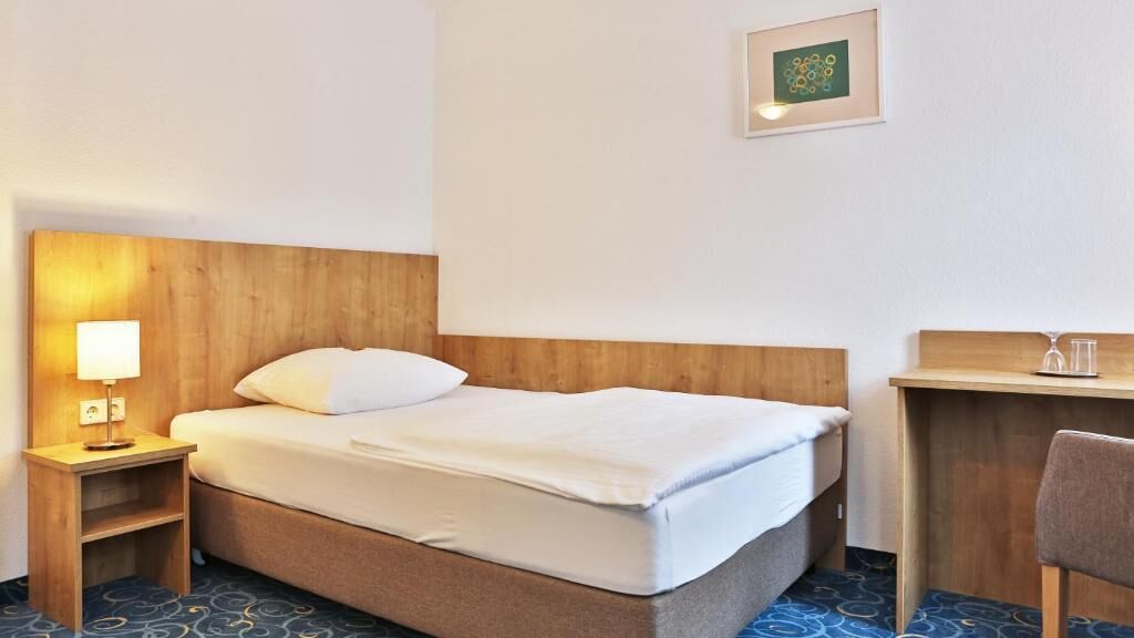 A hotel room with a wooden headboard, a single bed, a bedside table with a lamp, a small shelf, a desk with a chair, and a framed picture on the wall.