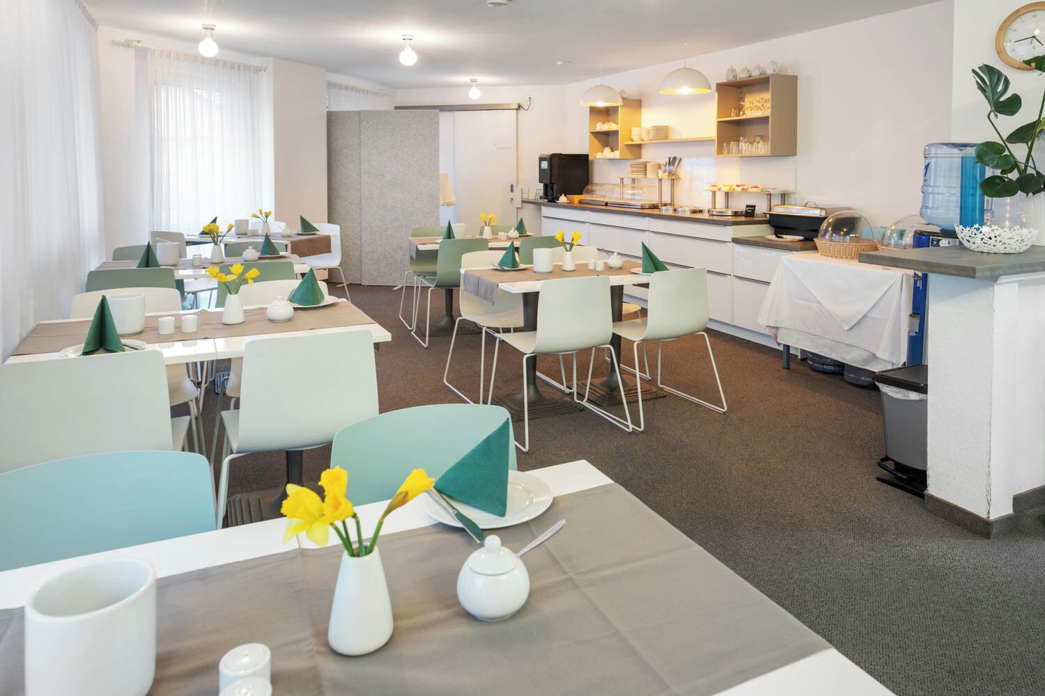 Ein moderner Hotel-Speisebereich mit gedeckten Tischen für das Frühstück, einer Küchenzeile und einem Wasserspender.