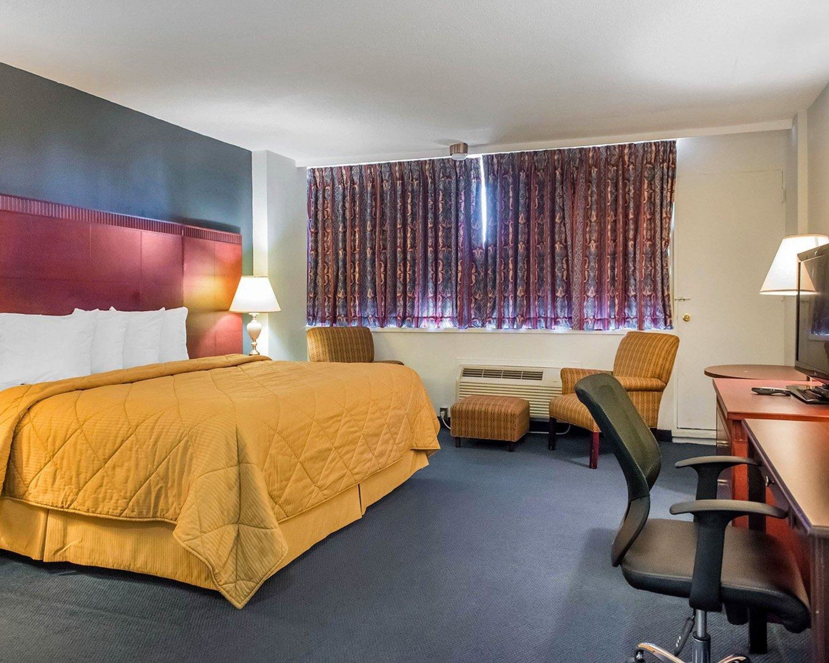 A hotel room with a large bed, a desk with a chair, a television, and a window with patterned curtains.