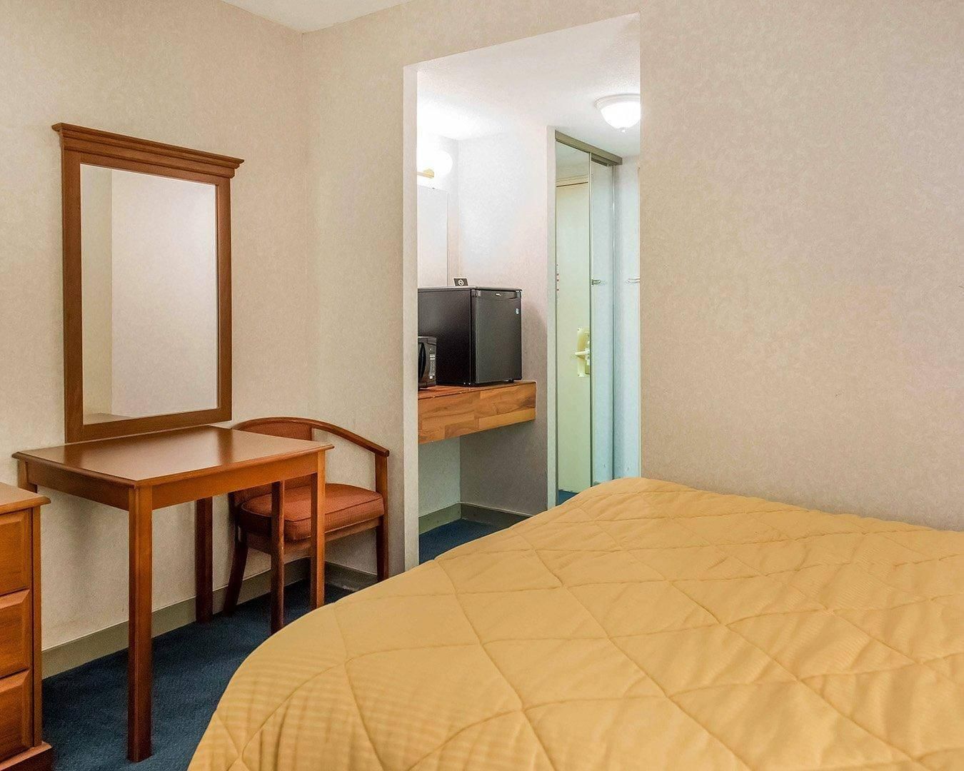 A hotel room with a bed, a desk with a chair, a mirror, a television, and a wardrobe.