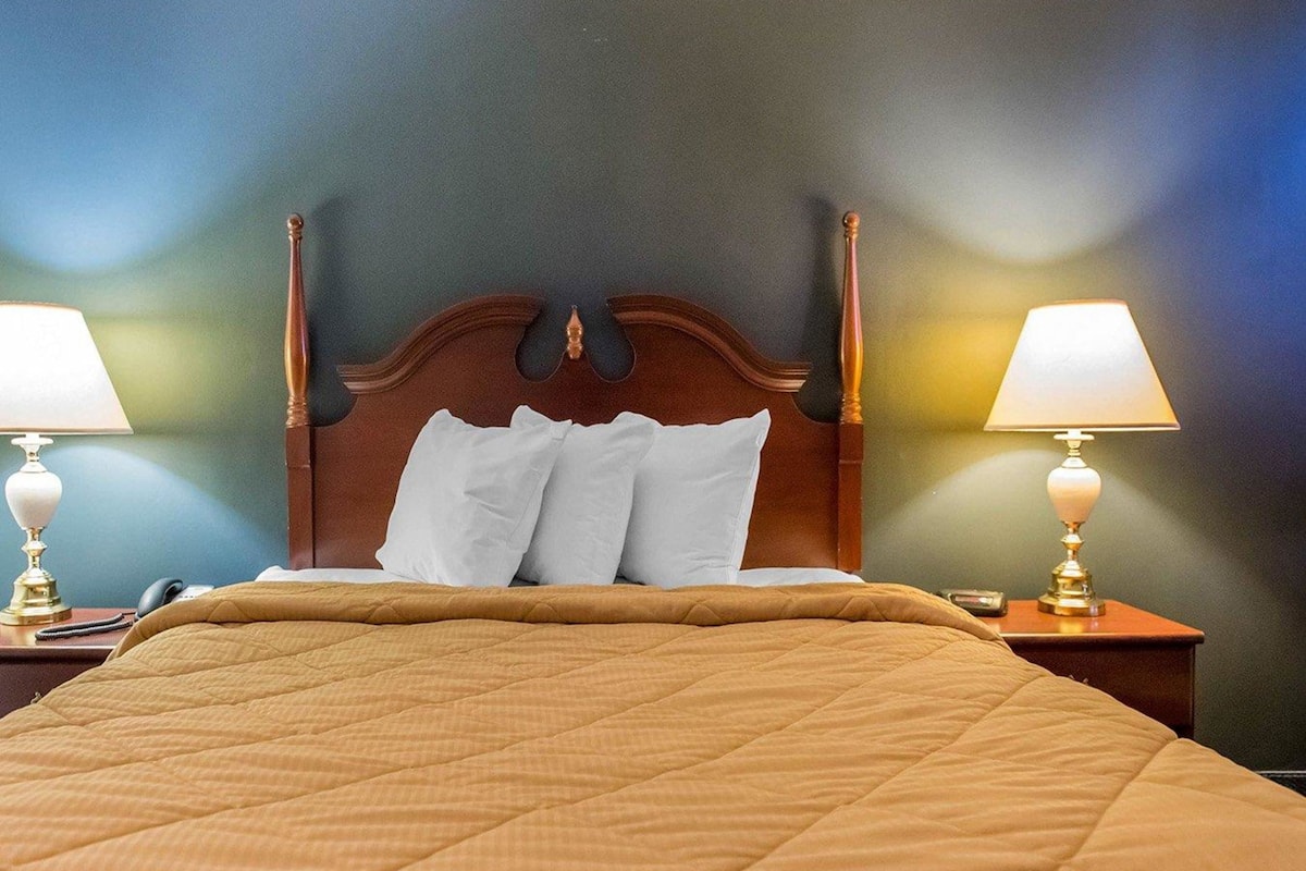 A hotel room with a large bed, wooden headboard, two bedside lamps, and a quilted bedspread.