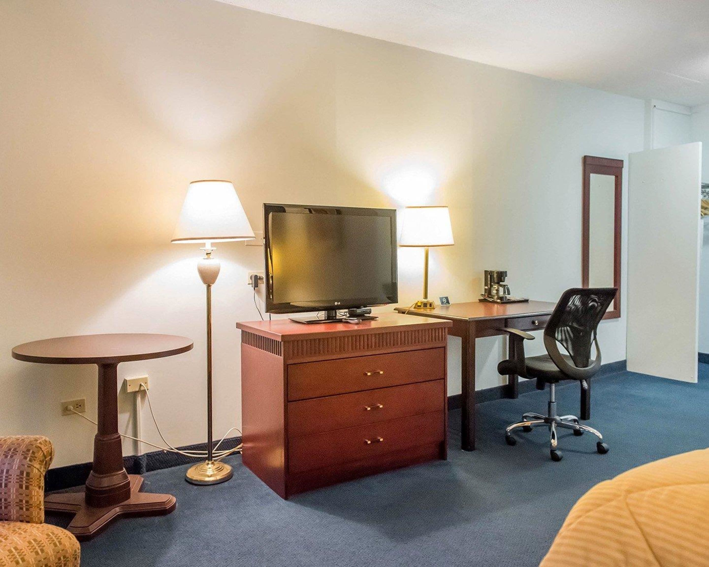 A hotel room with a desk, two lamps, a television, a coffee maker, a chair, and a dresser.