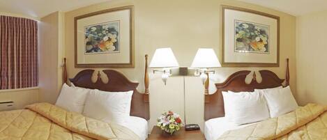 Habitación de hotel con dos camas, cada una con cabecero de madera, ropa de cama blanca y una mesita de noche con una lámpara y un florero.