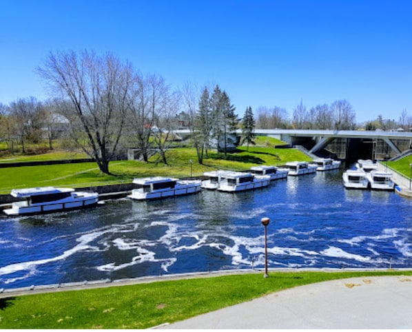 Une rivière bordée de bateaux amarrés, un pont en arrière-plan et une zone herbeuse avec des arbres.