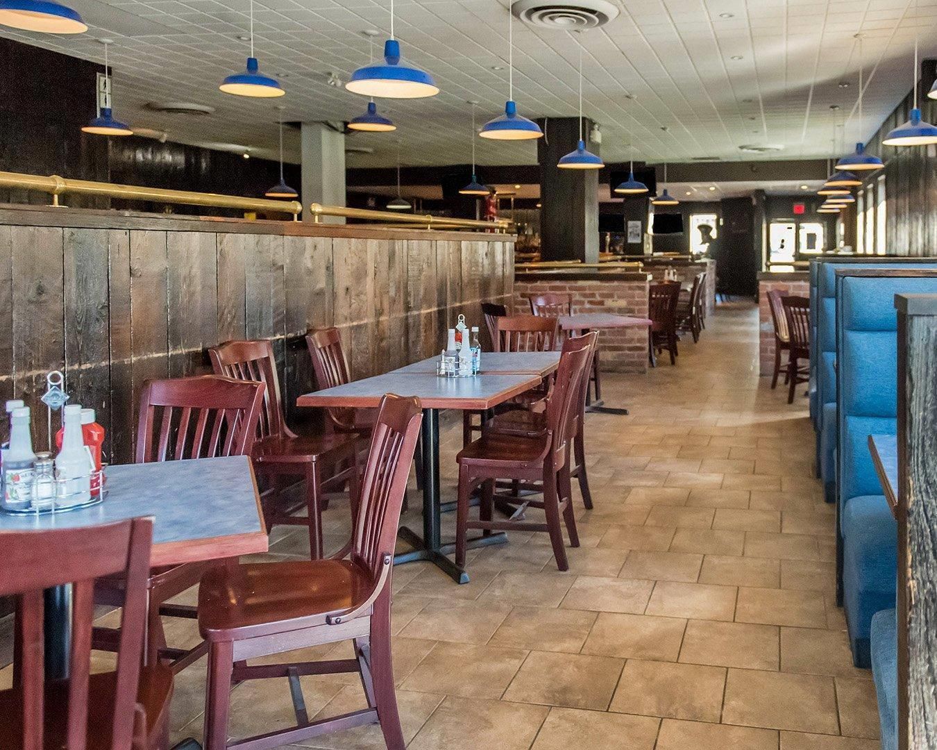 Un interior de restaurante con mesas y sillas de madera, lámparas colgantes azules y una pared de ladrillo.