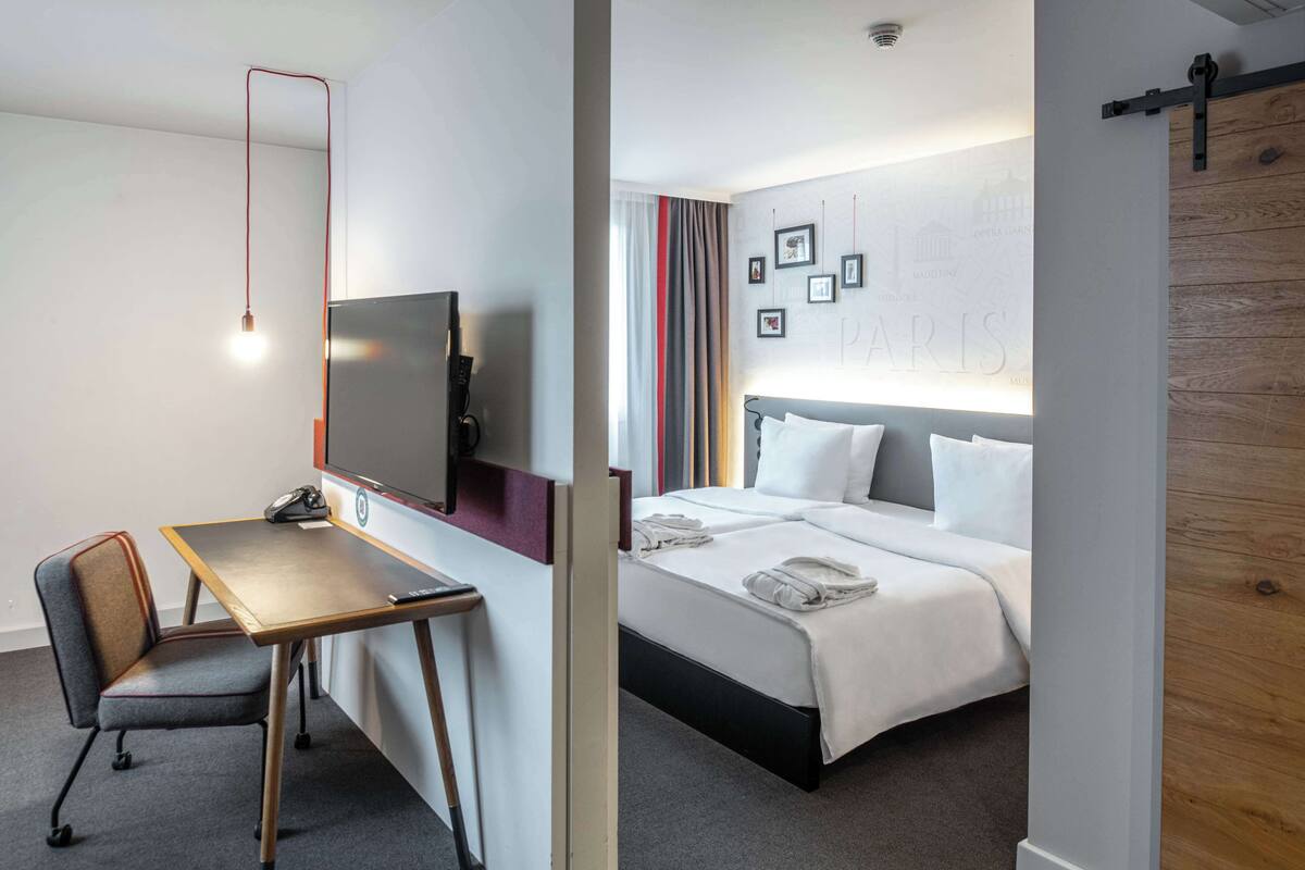 A hotel room with a bed, a desk, a chair, a TV, and a wall with framed pictures.
