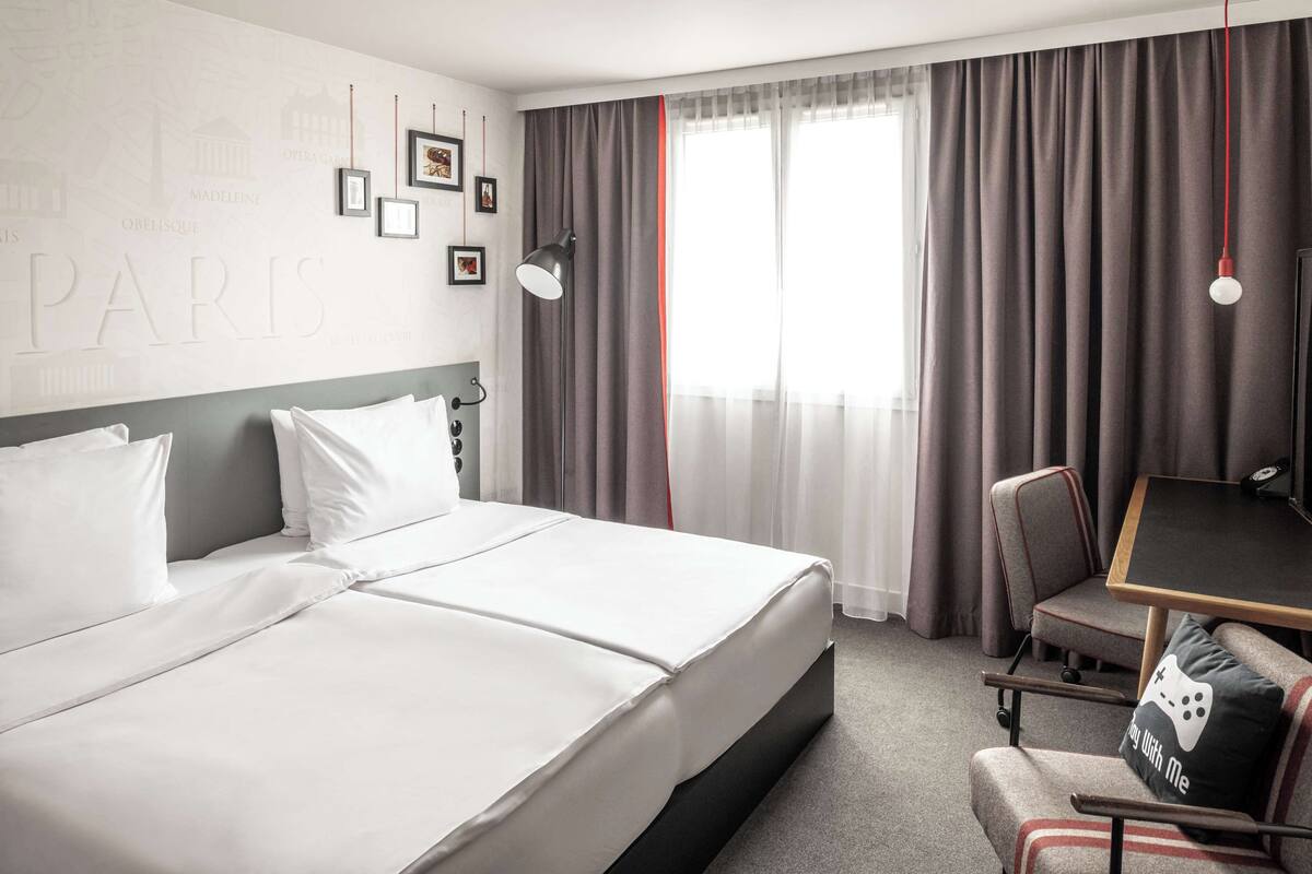 A hotel room with a large bed, a desk, and a chair. There are curtains on the window, and a wall with framed pictures.