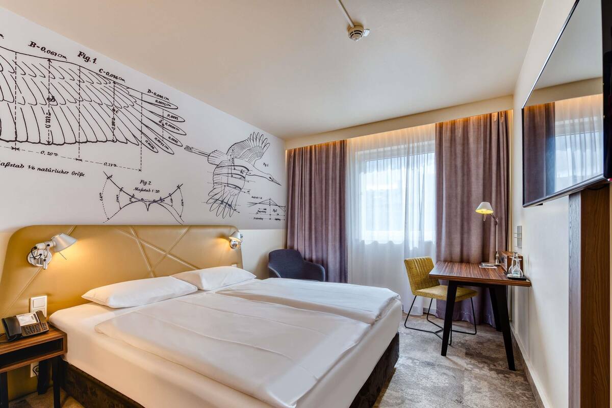 A hotel room with a large bed, a desk with a chair, a flat-screen TV, and a wall with diagrams.