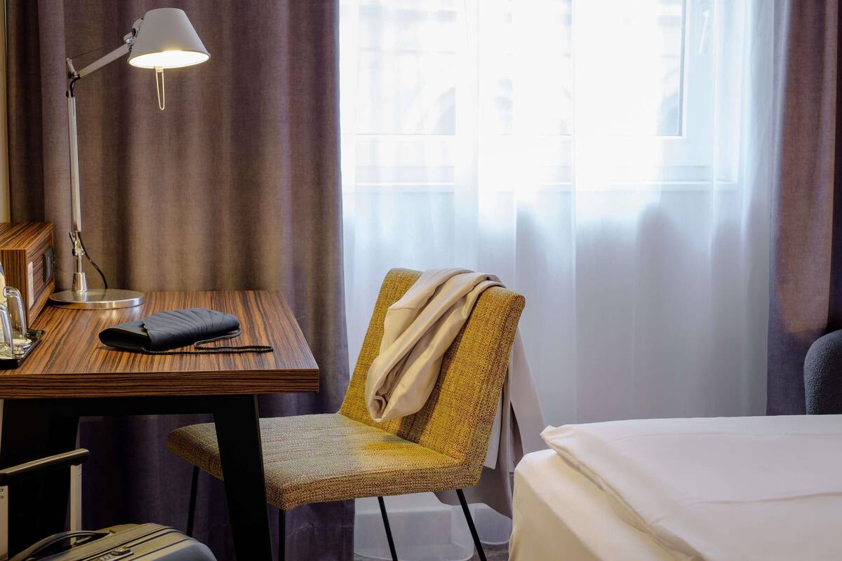 Una habitación de hotel con un escritorio de madera, una lámpara, una silla, una cama y una ventana con cortinas.