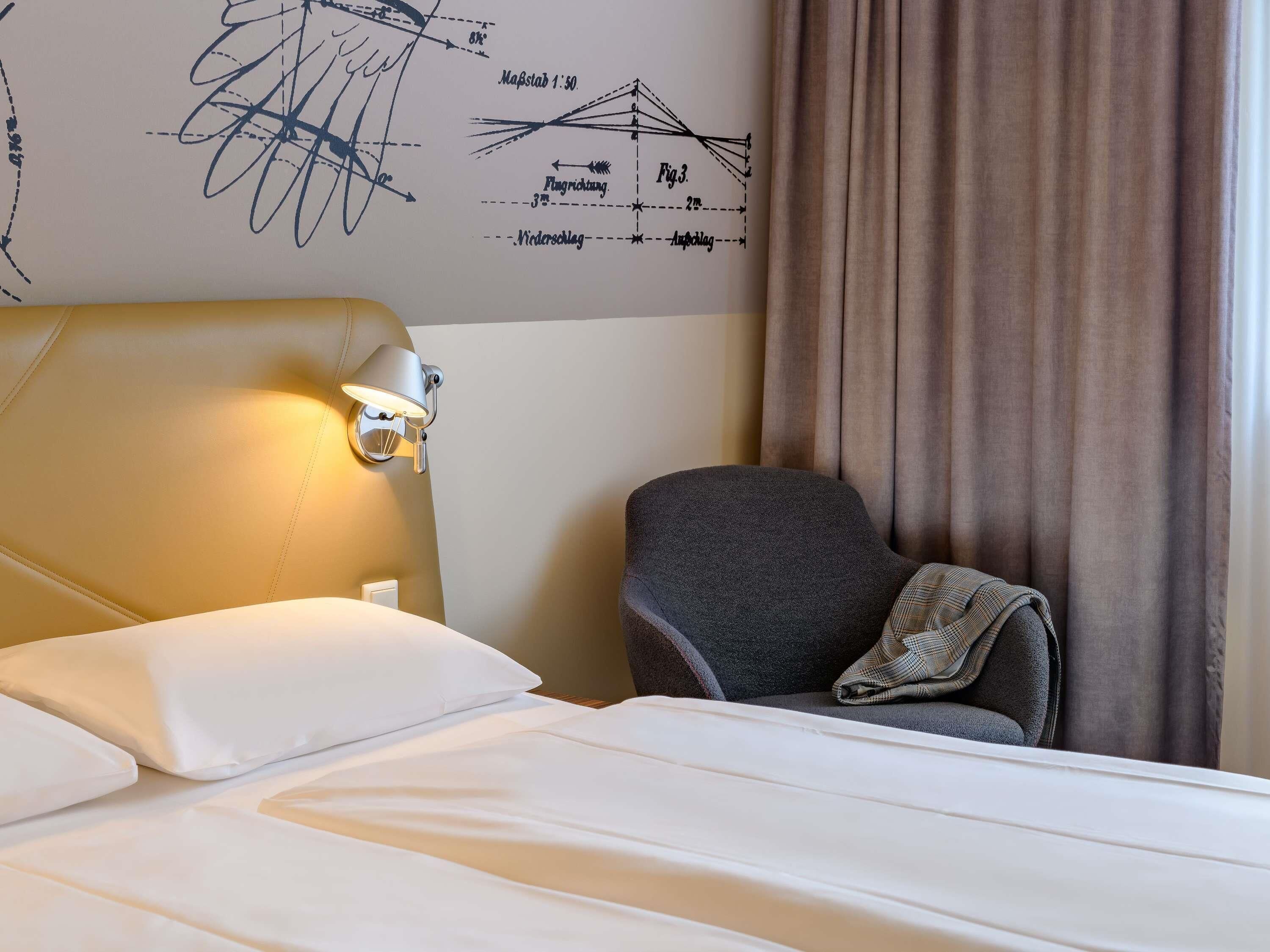 Habitación de hotel con cama, mesita de noche, lámpara, silla y una pared con un diagrama.