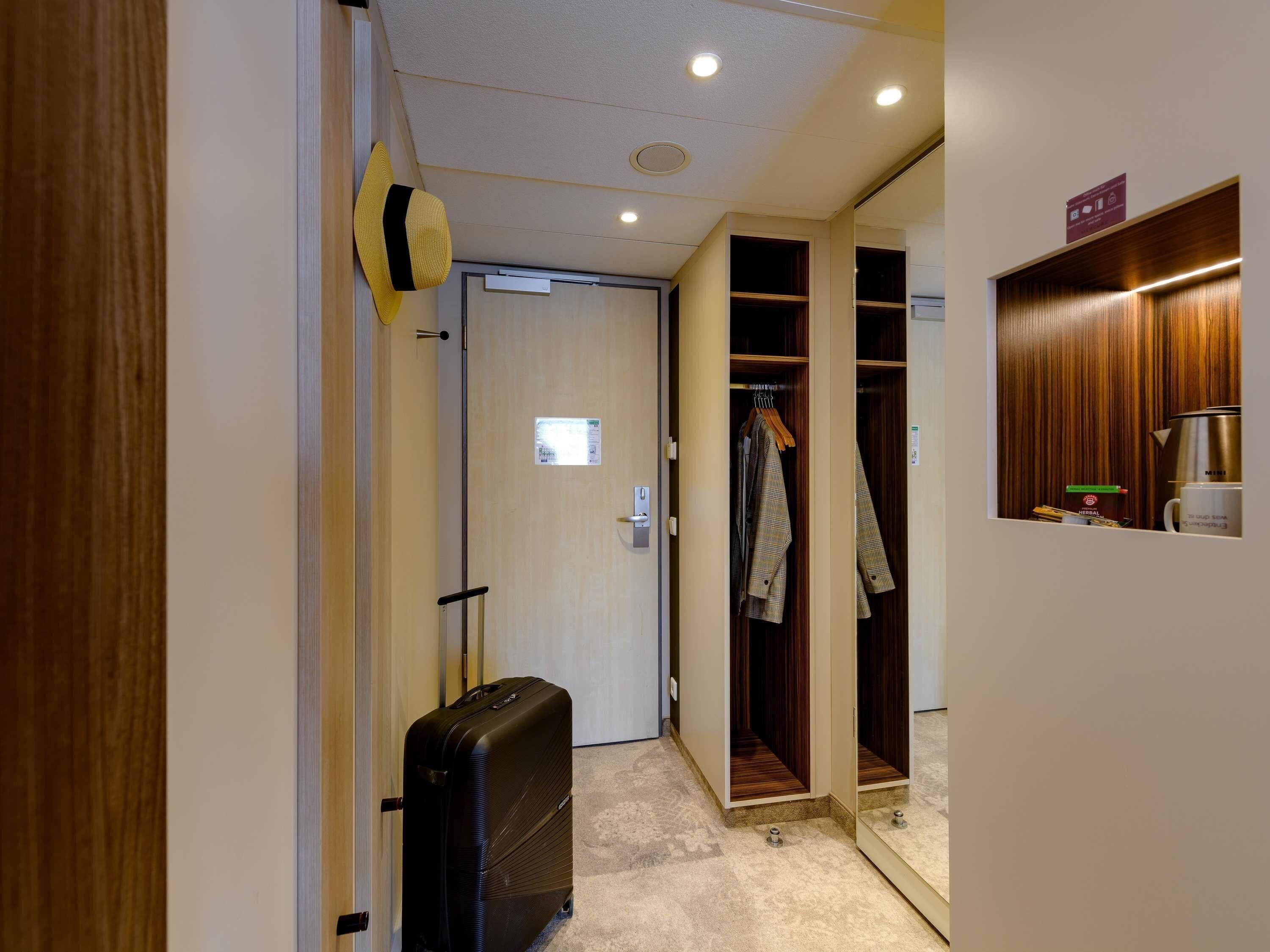 A hotel corridor with a wooden door, a black suitcase, a coat rack with a jacket, and a mirror reflecting the corridor.