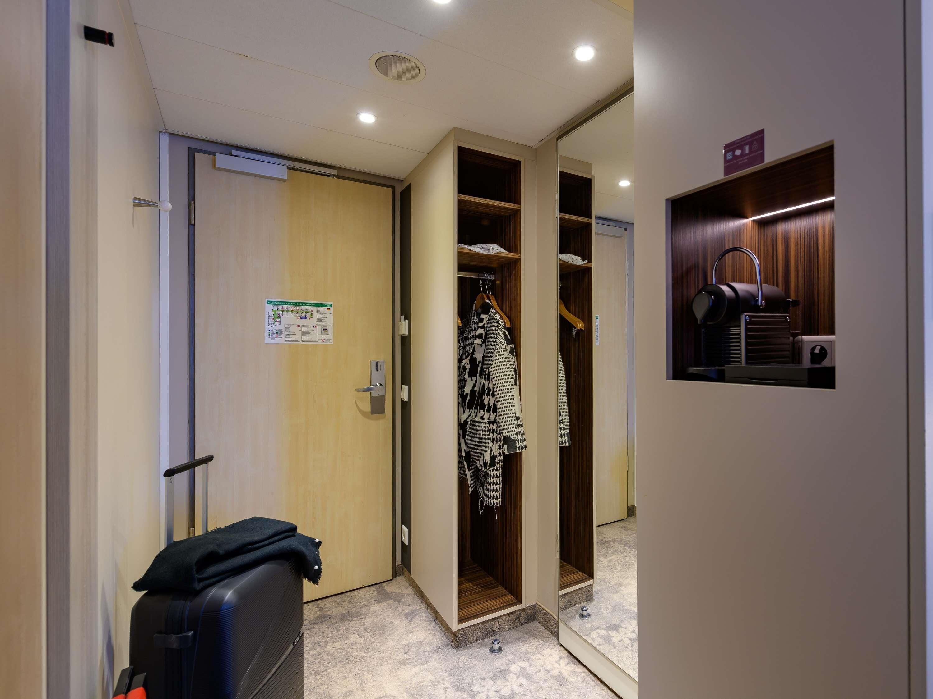 A hotel room with a wooden door, a coat rack with a patterned garment, a suitcase, and a small shelf with a speaker.