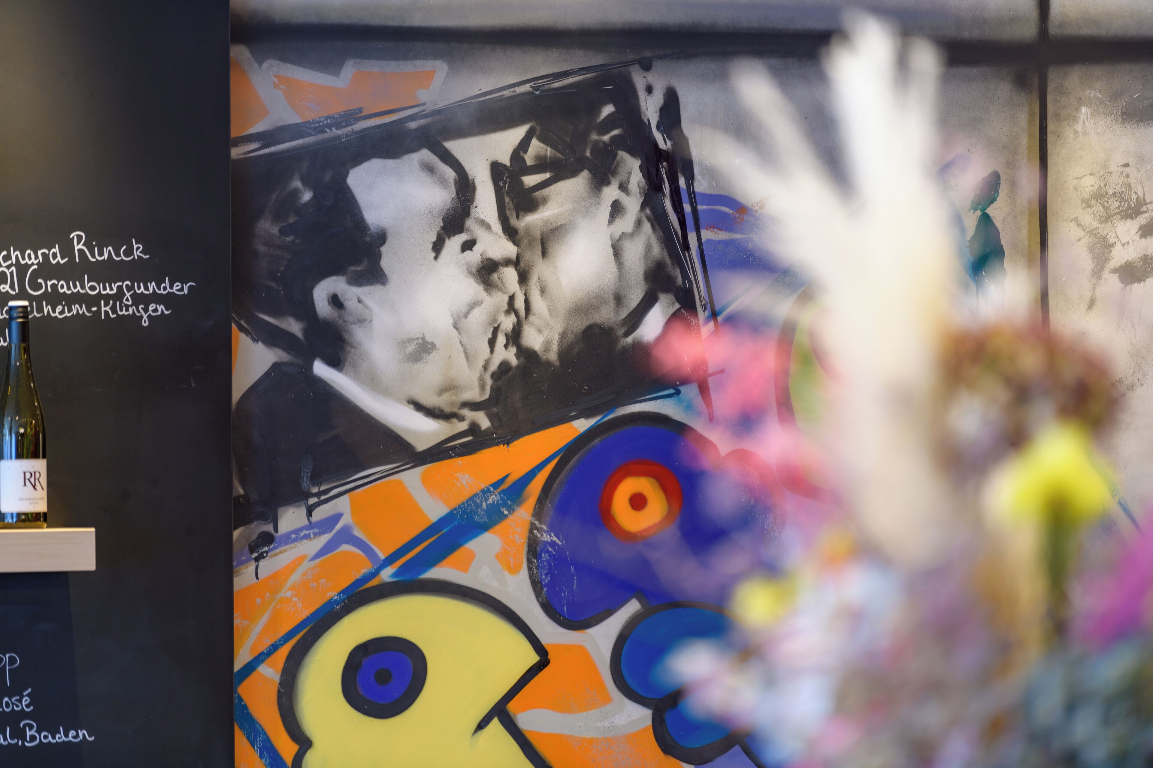 A wall with a graffiti-style artwork, a bottle of wine, and a floral arrangement.