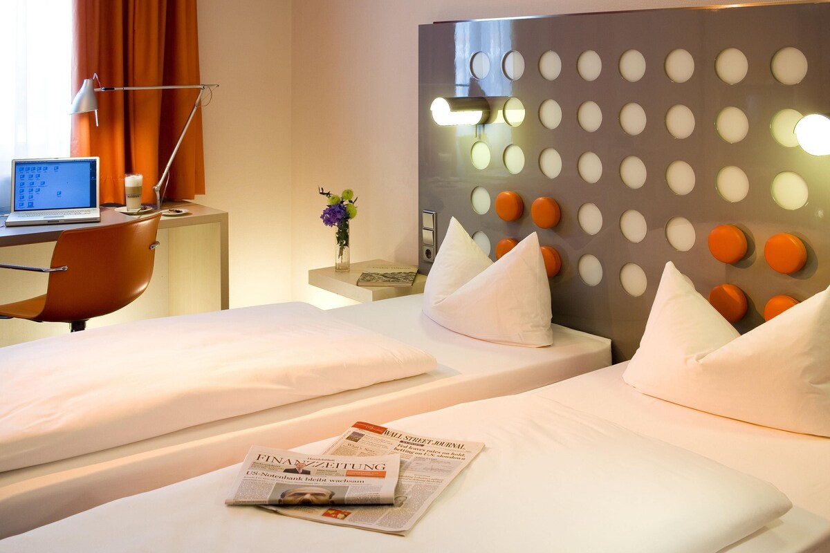 A hotel room with two beds, a desk with a laptop, a vase with flowers, and a wall with circular light fixtures.