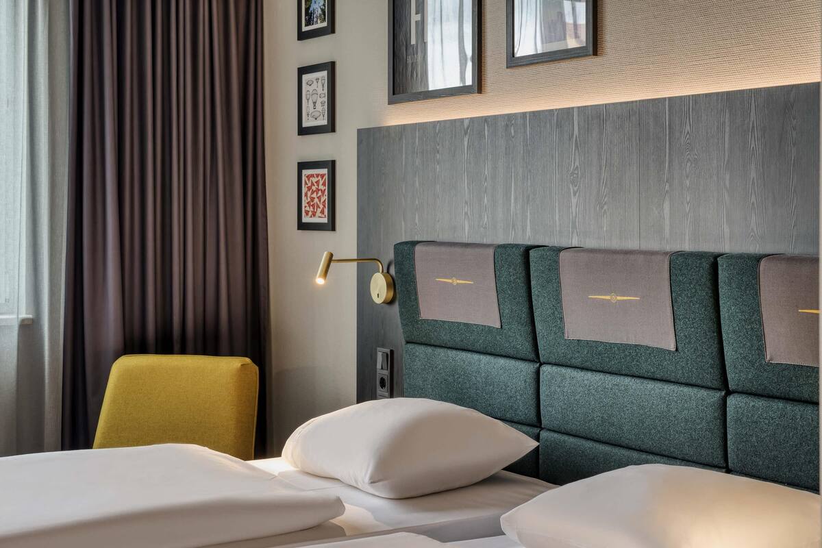 A hotel room with a bed, a yellow chair, a wall-mounted lamp, and framed pictures.