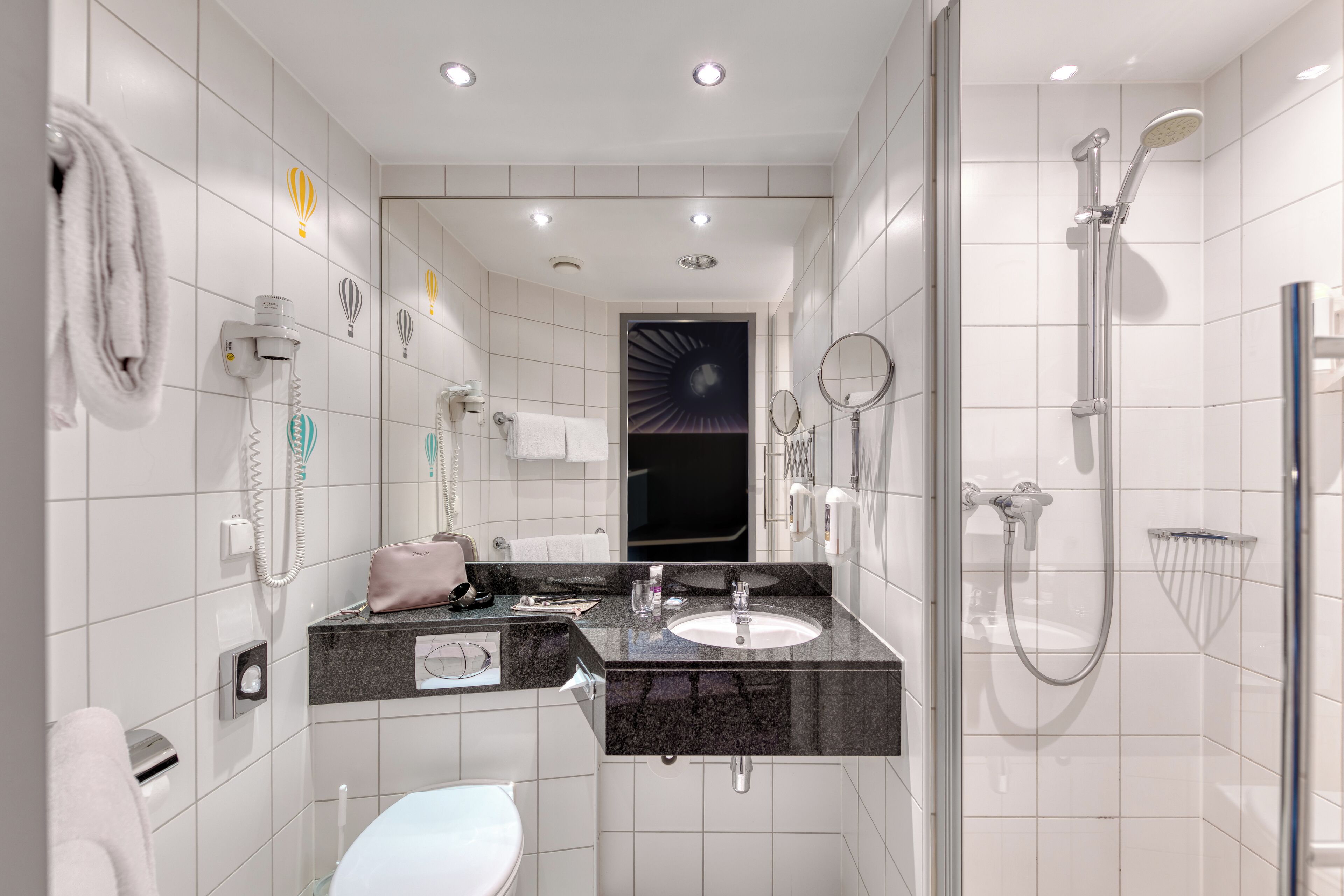 A modern bathroom with a black granite countertop, white tiles, a glass-enclosed shower, and a visible towel rack.