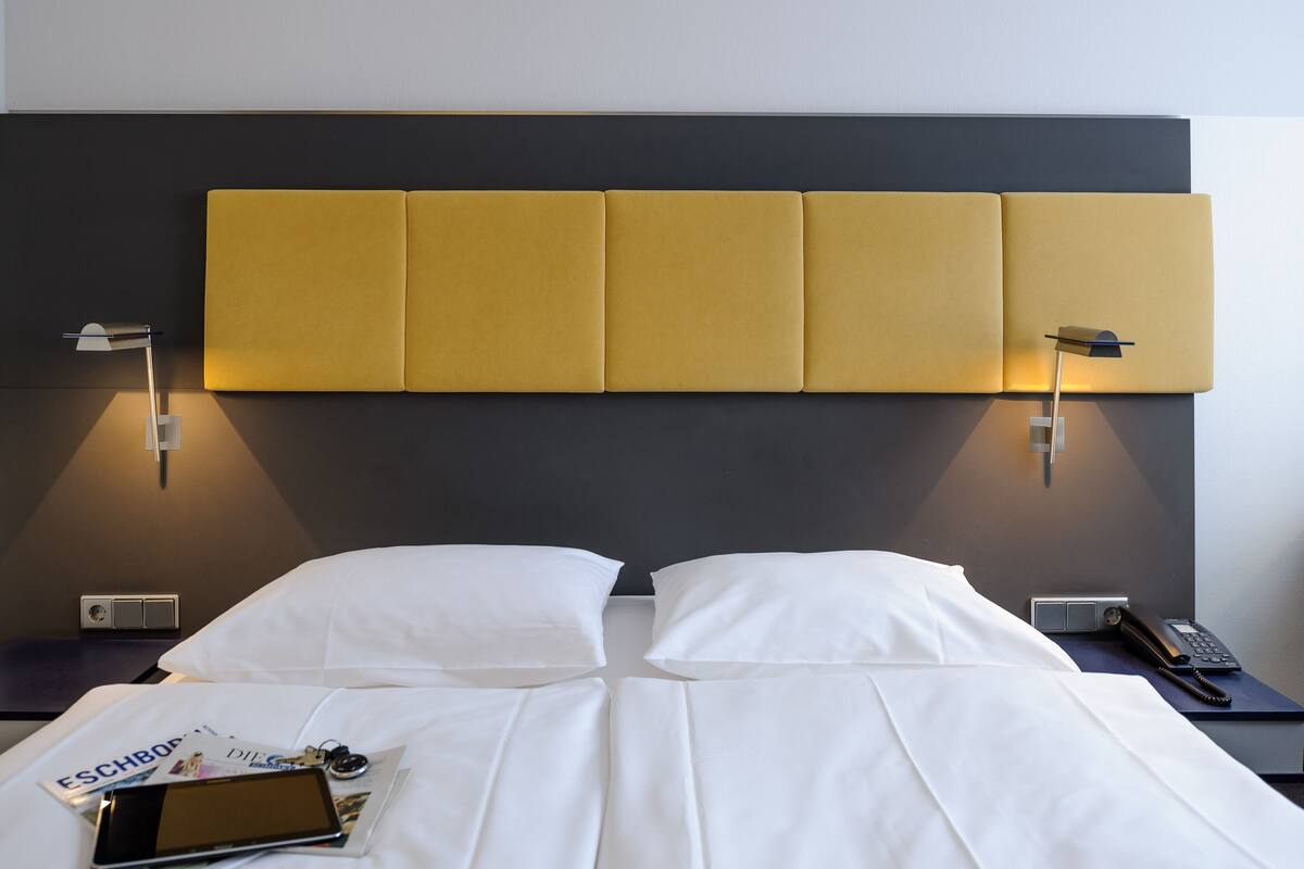 Une chambre d’hôtel au design moderne, dotée d’un grand lit avec du linge de lit blanc, d’une tête de lit ornée d’un panneau jaune original, de tables de chevet avec des lampes et d’un téléphone sur la table de chevet.
