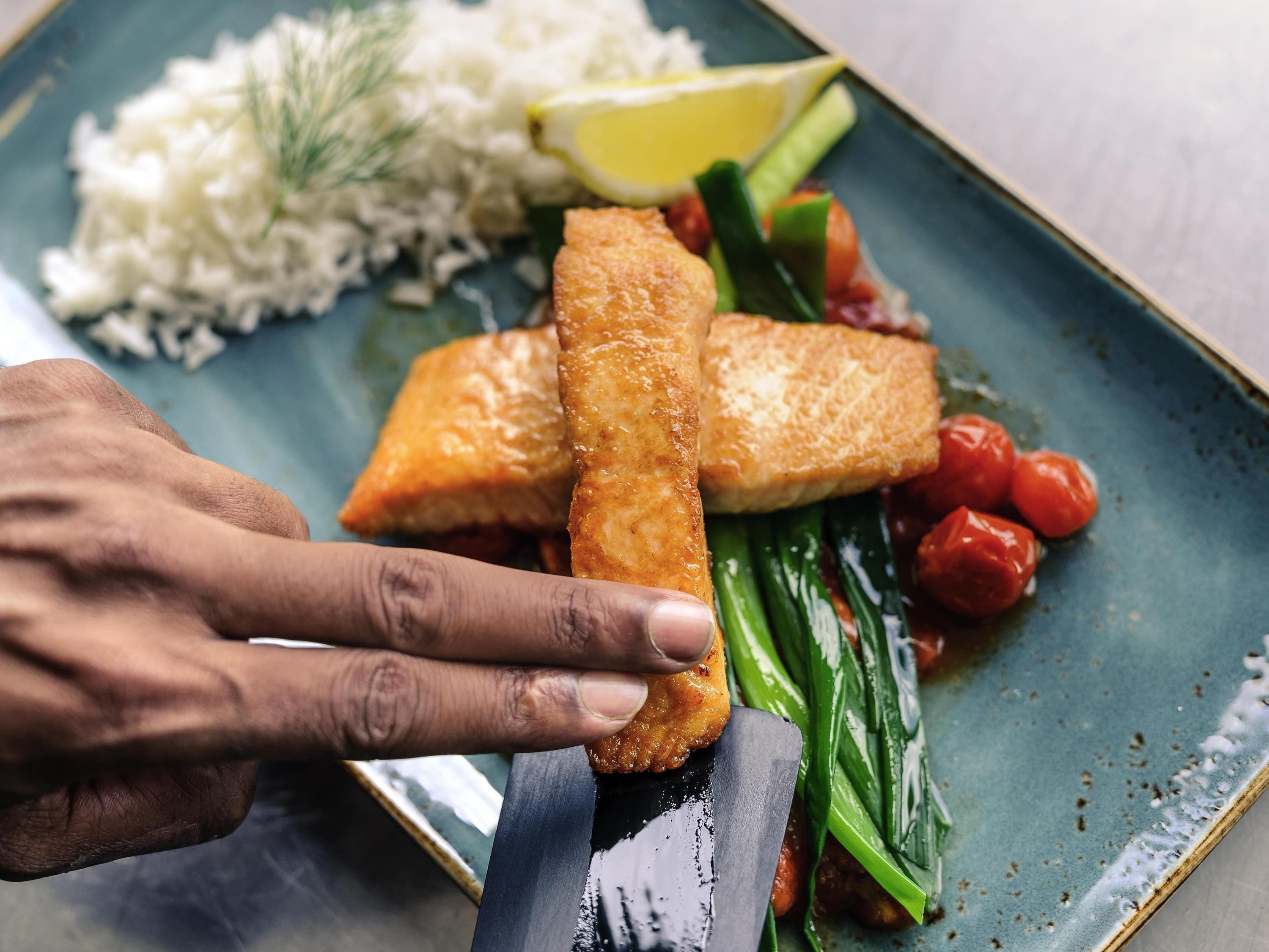 Una mano sosteniendo un cuchillo, cortando un alimento empanizado sobre un plato con arroz, verduras y un gajo de limón.